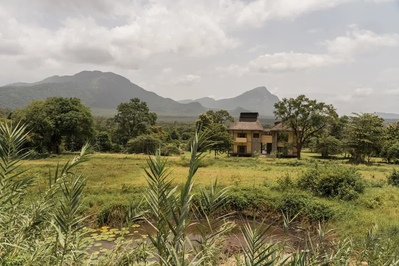 Two-storey residence set in a grassy clearing with mountains and scattered trees under cloudy skies