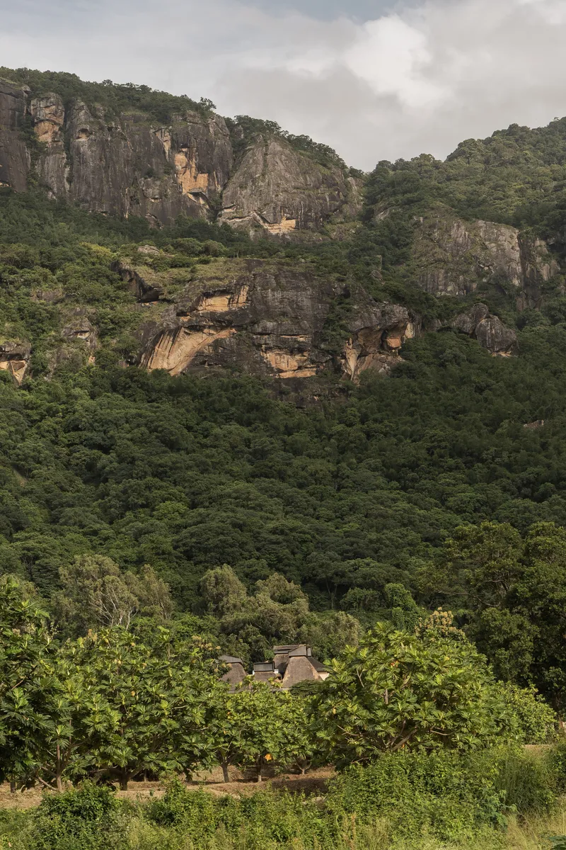 Distant view of low-rise structures nestled in dense vegetation beneath layered rock cliffs under cloudy skies