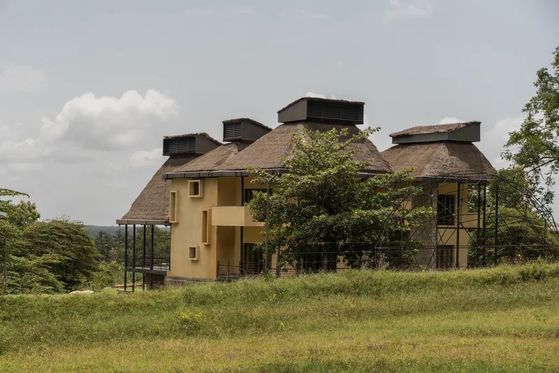 Yellow plastered volumes with thatched roofs and ventilation towers set in grassland under cloudy skies