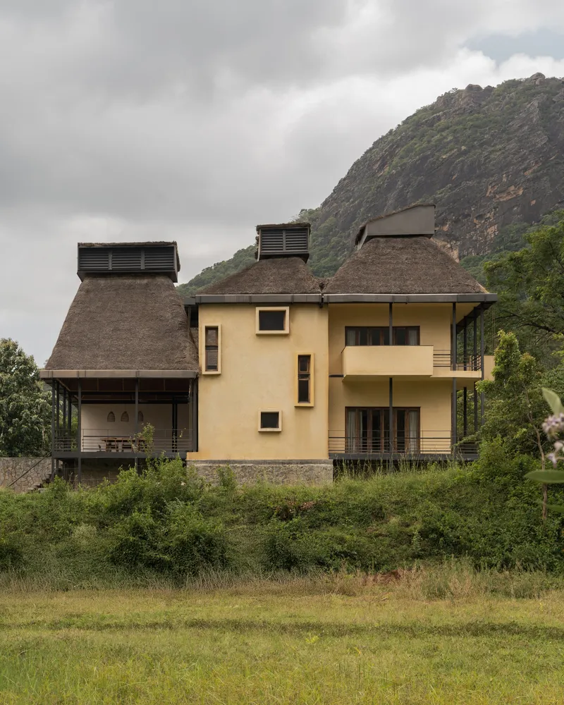 Facade view showing thatched roof pavilions with ventilation towers against a mountain backdrop in overcast weather