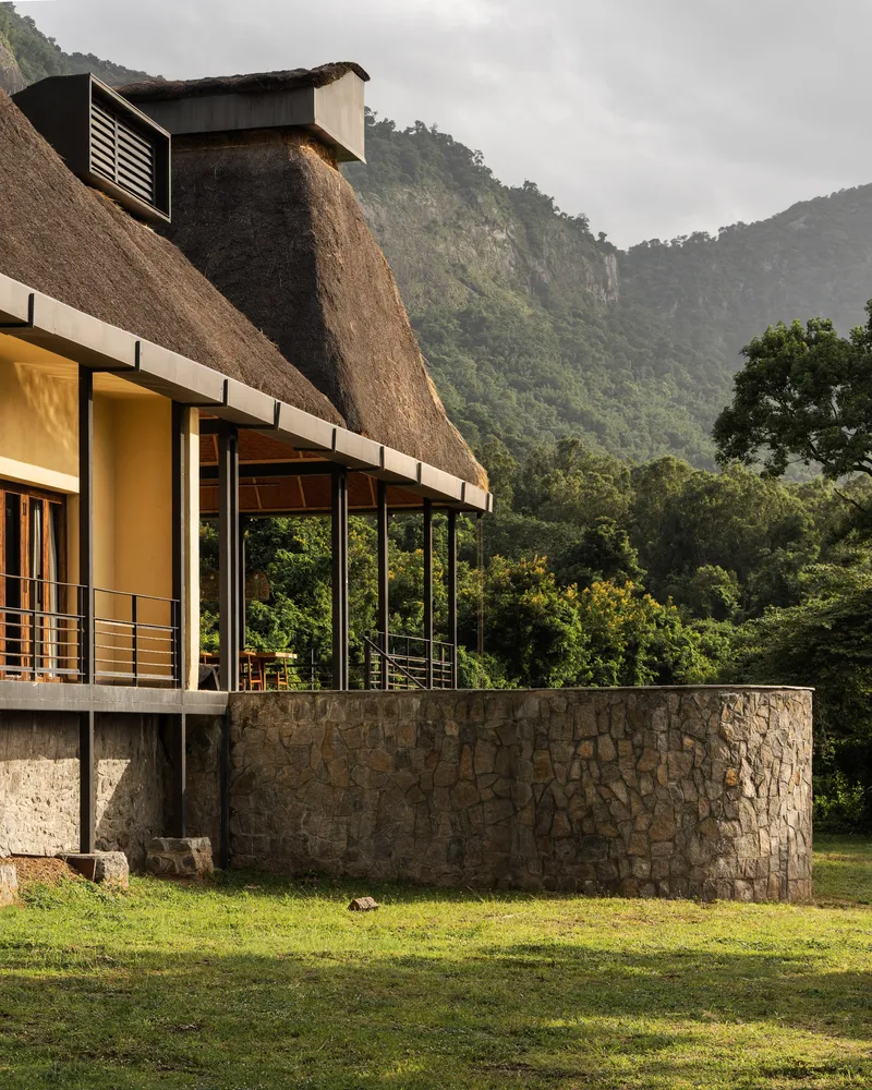 Curved stone retaining wall at base of a thatched roof structure with forested mountains in the background