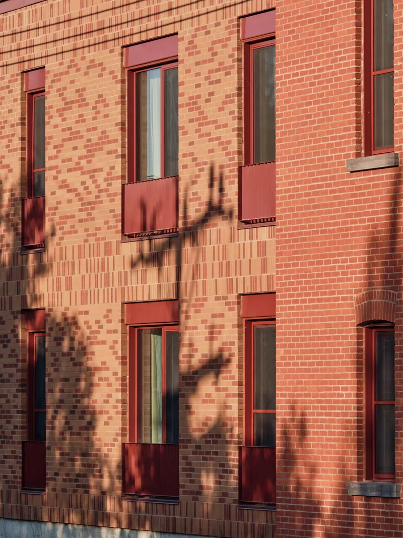 Facade detail showing patterned brickwork with tree shadows cast across windows and red metal panels