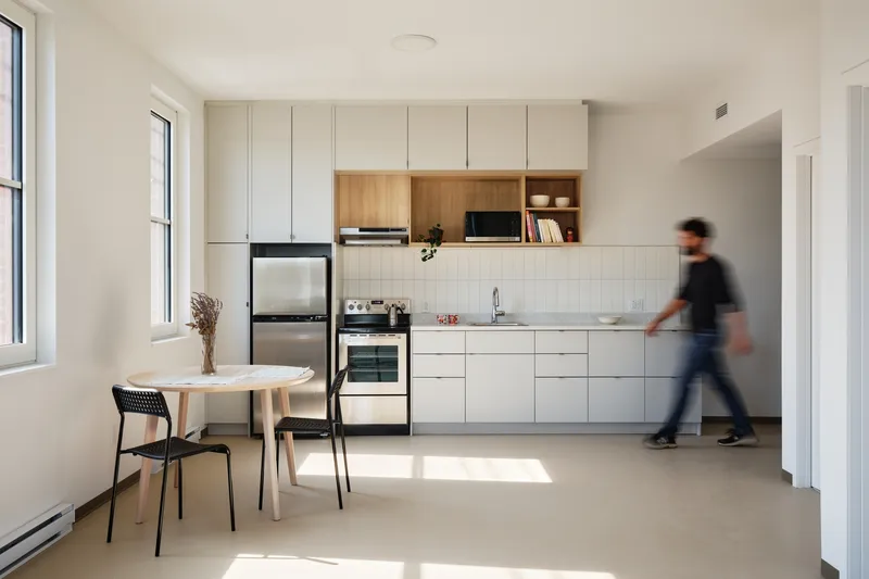 Kitchenette with a figure in motion blur walking past sunlit windows and pale wood cabinetry