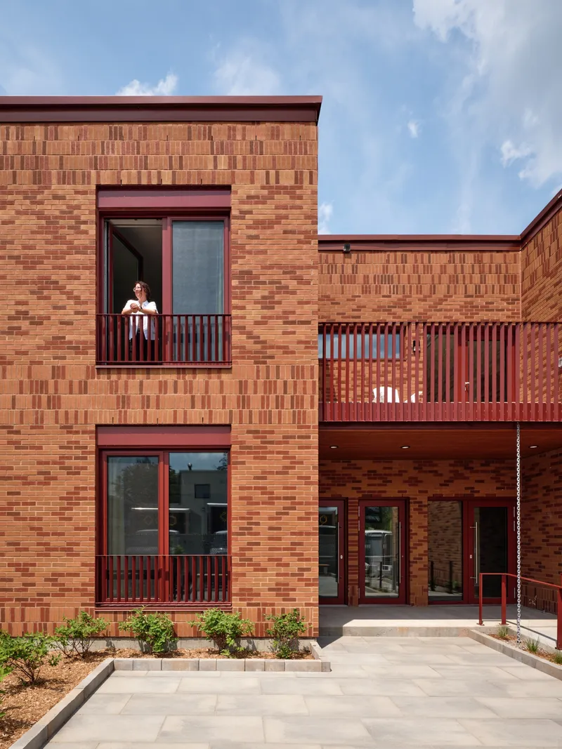 Brick facade with stacked balconies and red metal railings where a resident stands in afternoon sun