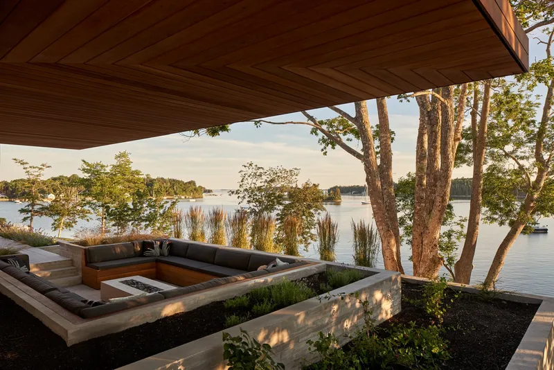 Covered terrace with sunken seating and fire pit overlooking a lake through mature trees