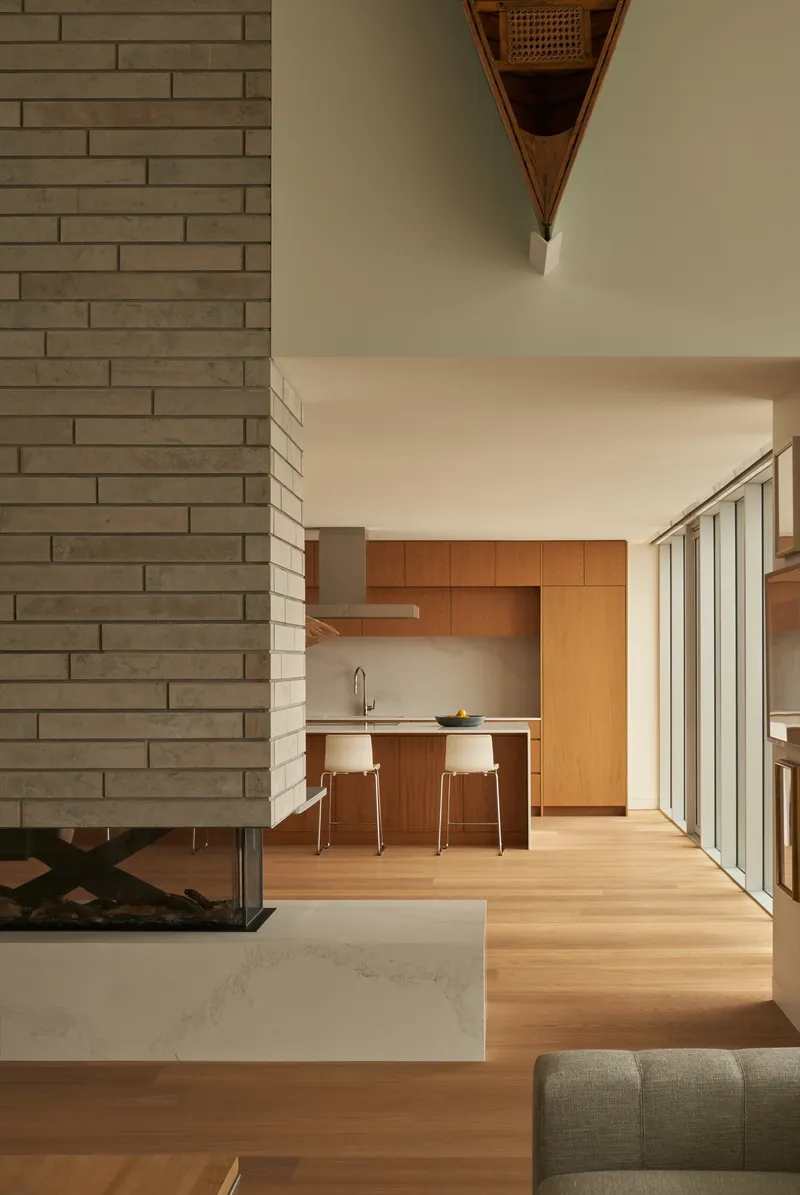 Open-plan interior showing the brick fireplace volume and kitchen with timber cabinetry under soft afternoon light