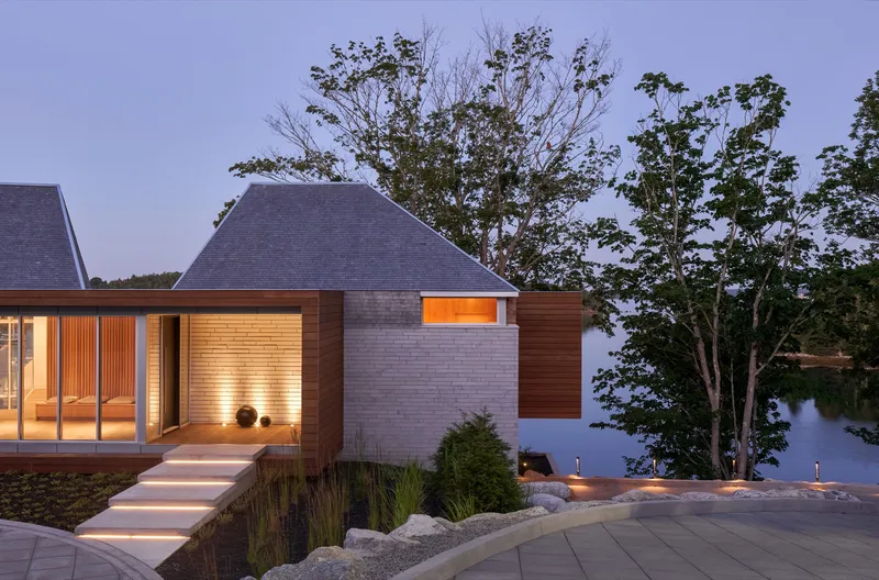 Illuminated pavilion volumes with brick and timber cladding beside a lake at twilight