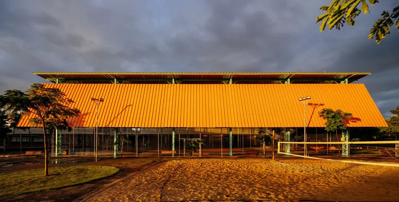 Glazed pavilion with an orange corrugated metal roof illuminated by golden hour sunlight under stormy clouds