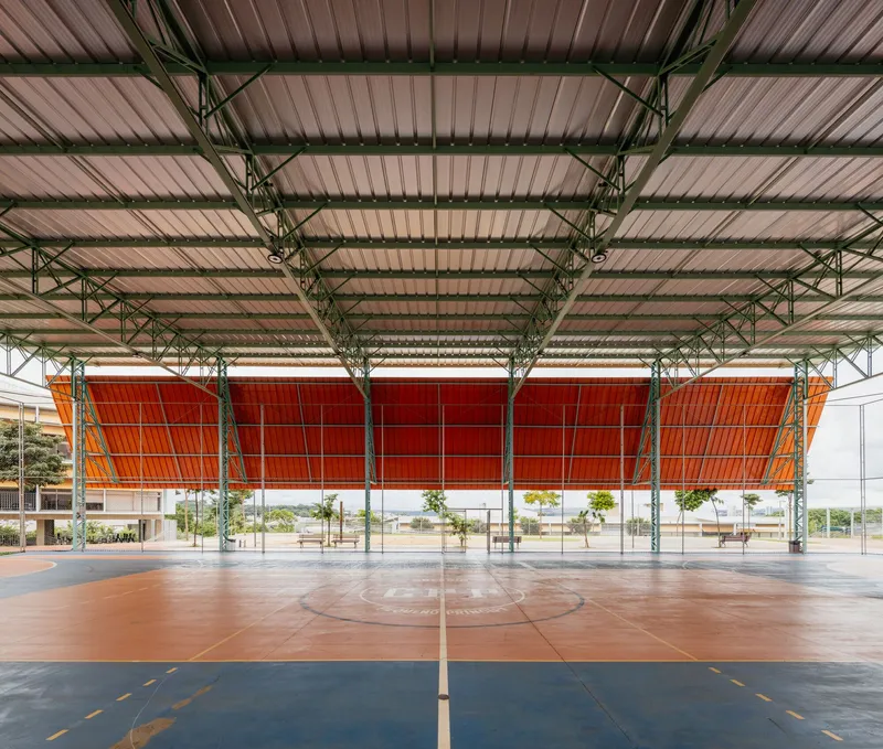 Covered sports court with green steel trusses and orange perforated wall panels behind planted trees