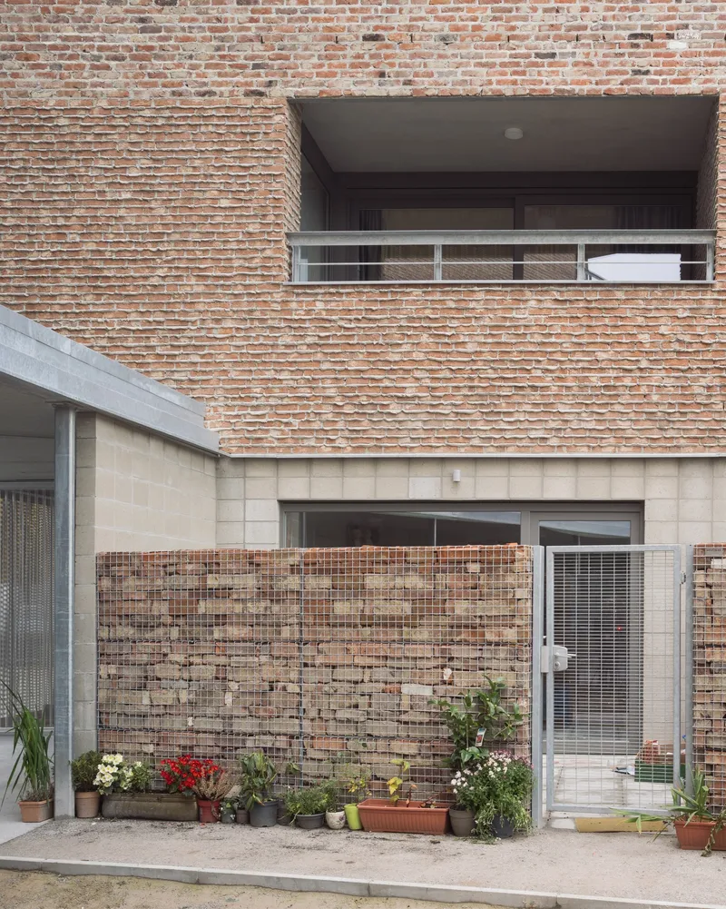 Ground floor courtyard with gabion wall and potted plants beneath cantilevered brick facade and balcony