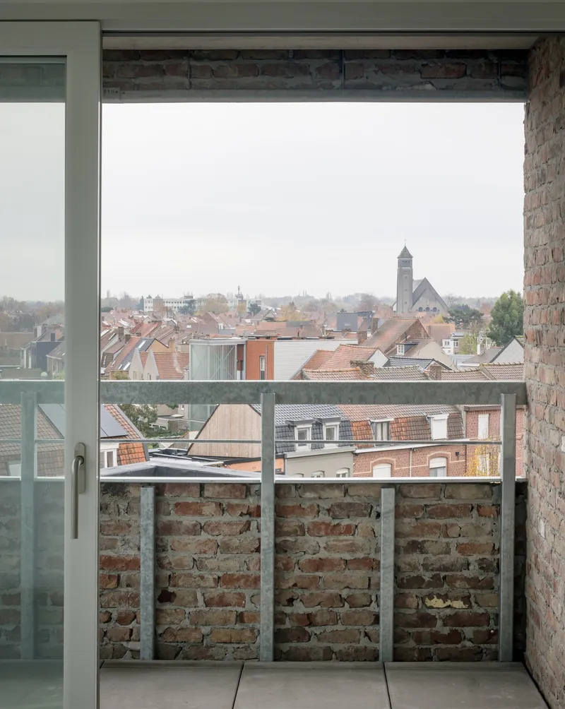 View from a balcony through steel-framed glass doors overlooking red-tiled rooftops and a distant church spire