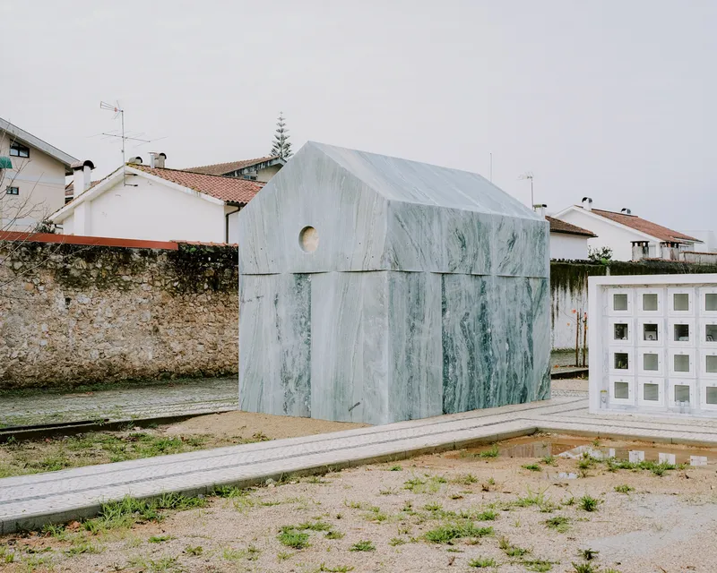 Gable-roofed pavilion clad in veined green marble panels beside a paved plaza under overcast skies