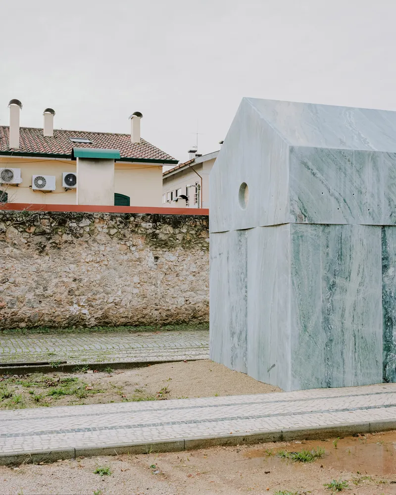 Side elevation showing layered marble panels against a weathered stone boundary wall and neighboring houses