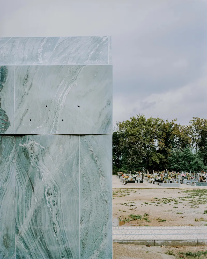 Corner of the marble chapel with cemetery grounds and trees visible beyond under cloudy sky