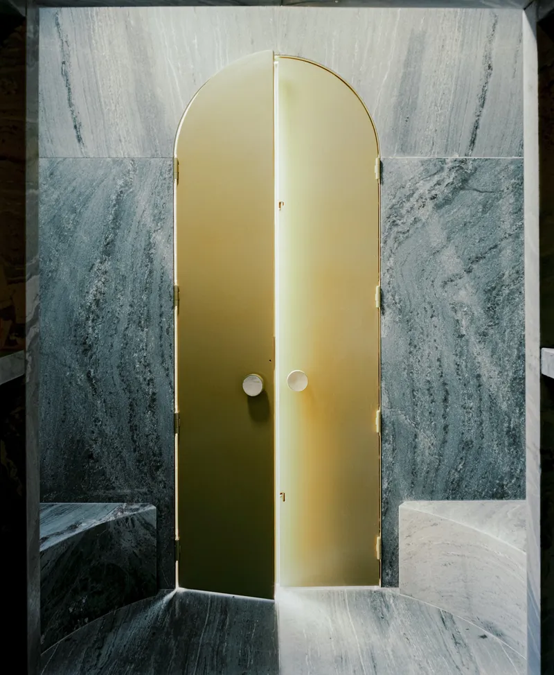 Arched brass doors set within a veined grey marble wall with recessed lighting along the edges