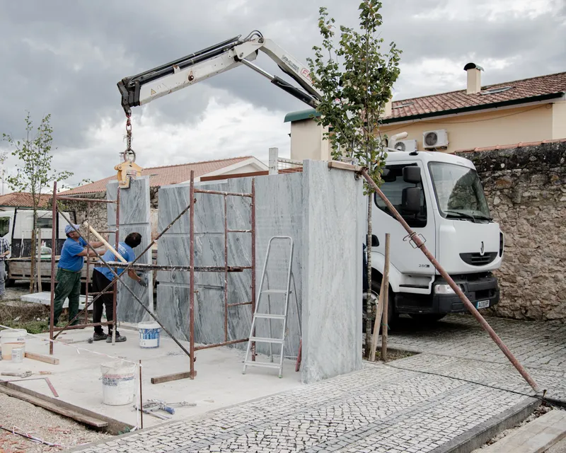 Construction workers installing prefabricated marble-clad panels with a crane-mounted truck beside young trees