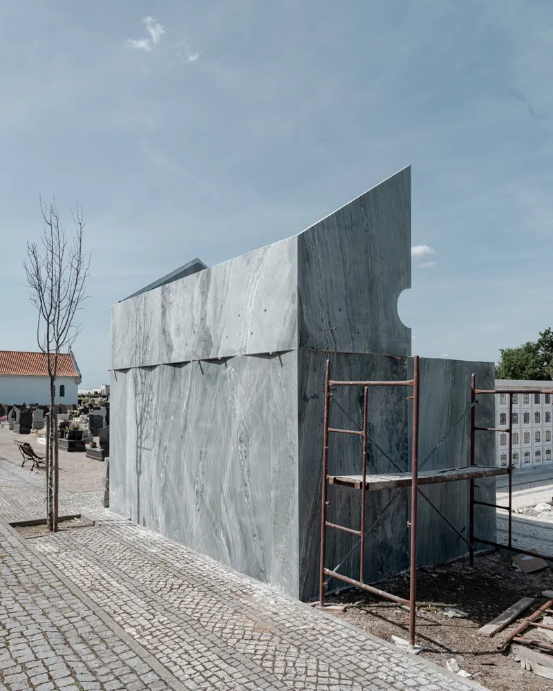 Exterior view of the angled marble-clad facade with metal scaffolding and a cemetery in the background