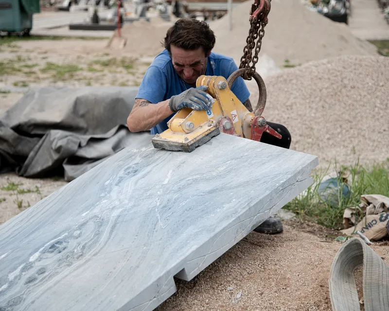 Worker cutting a large grey marble slab with a chain saw in an outdoor construction yard