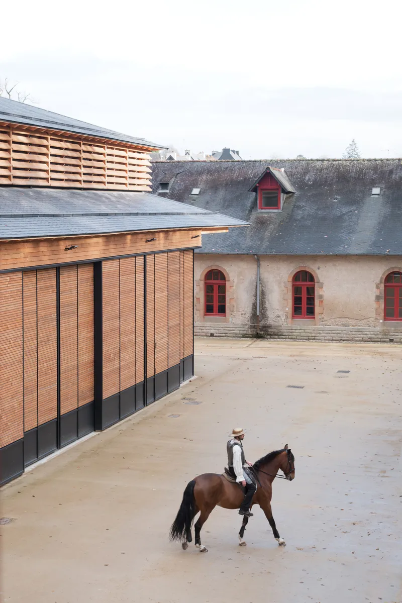 Courtyard view of the horizontal slatted timber facade beside a weathered stone stable with a rider on horseback