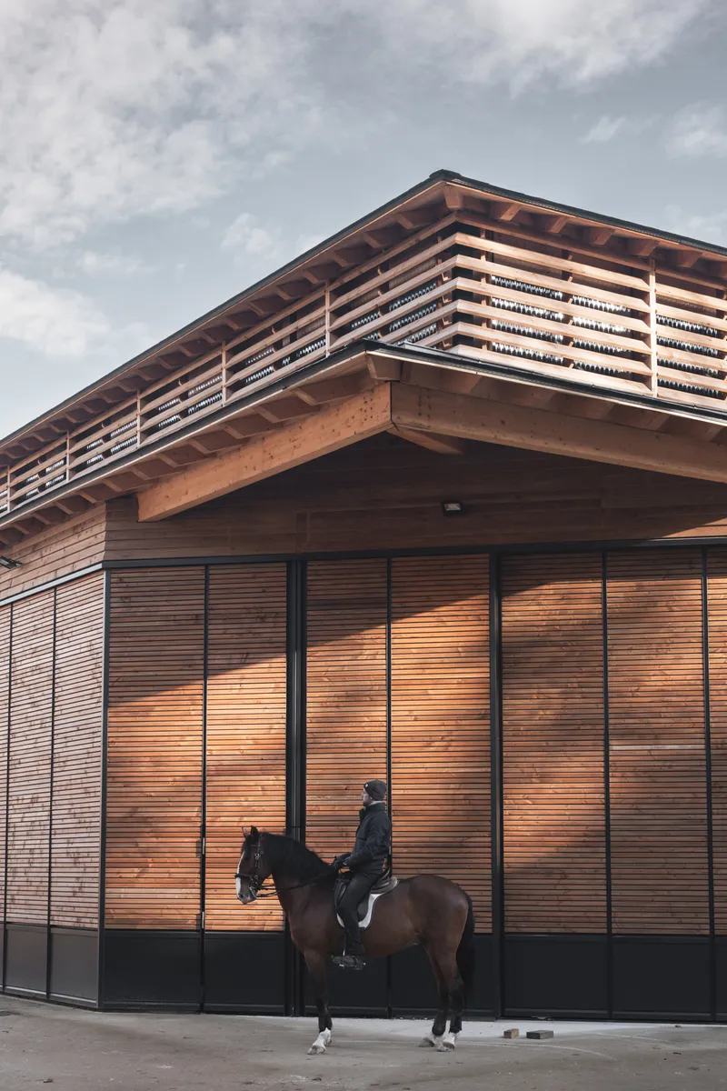 Corner facade of horizontal timber slats with exposed roof rafters and a rider on horseback below