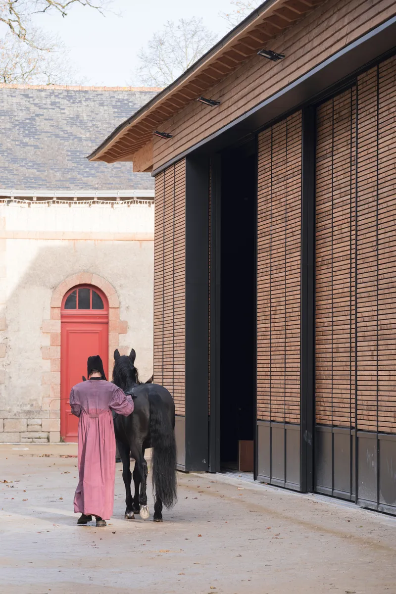 Person leading a black horse past the slatted timber wall and stone building entrance in morning light