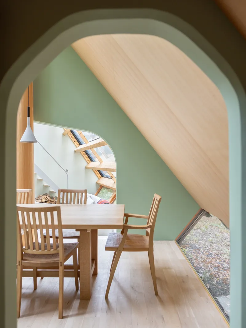 View through pale green arch to dining area with timber table and white staircase beneath sloping plywood ceiling