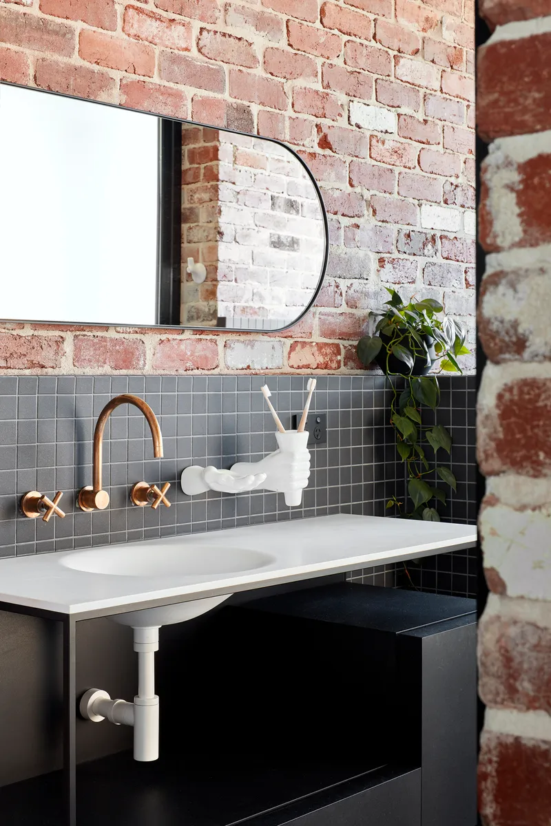 Bathroom vanity with grey mosaic tile backsplash and wall-mounted brass fixtures below exposed brick