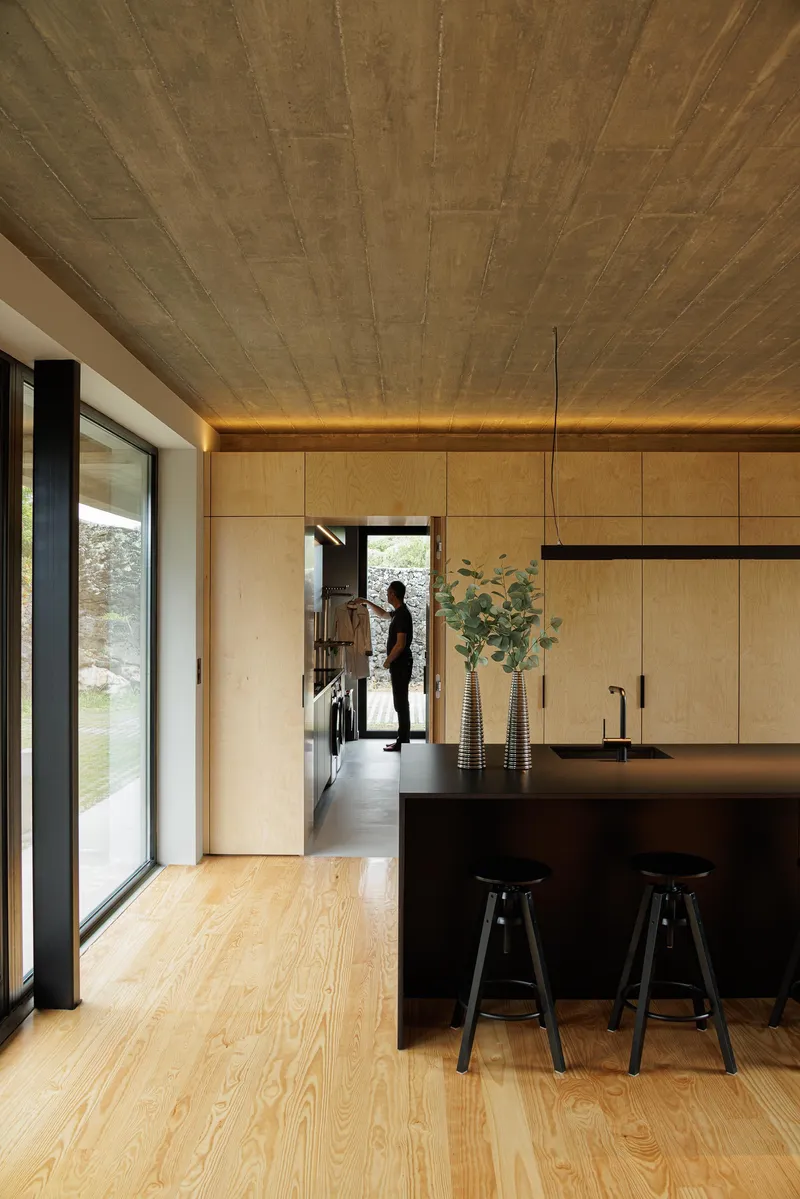 Kitchen with black island and plywood cabinetry beneath an exposed concrete ceiling with figure silhouetted in doorway