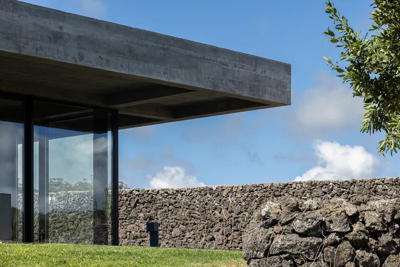 Cantilevered concrete roof slab with exposed formwork soffit above a glazed volume and volcanic stone wall