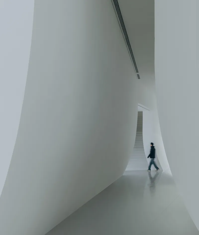 White interior corridor with a person walking beneath curving walls and a linear ceiling slot