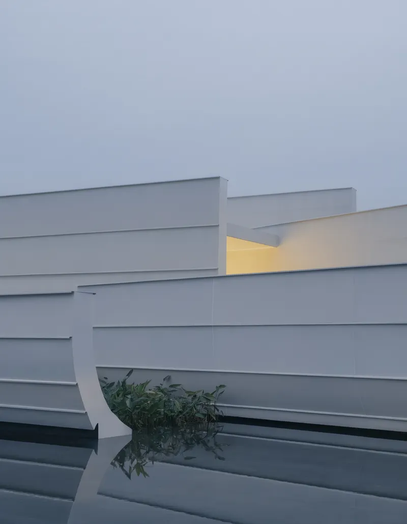 Layered white horizontal bands with illuminated recess and curved reflecting pool with greenery at twilight