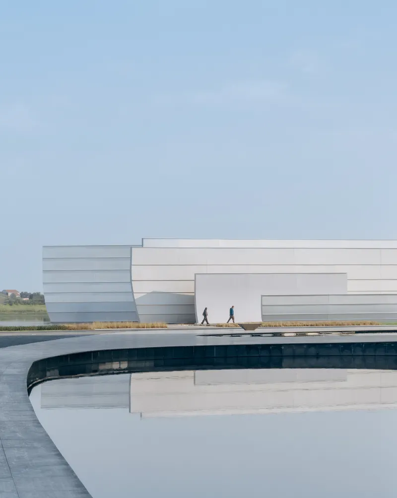 Curved water channel beside the white layered facade with two figures walking on the plaza