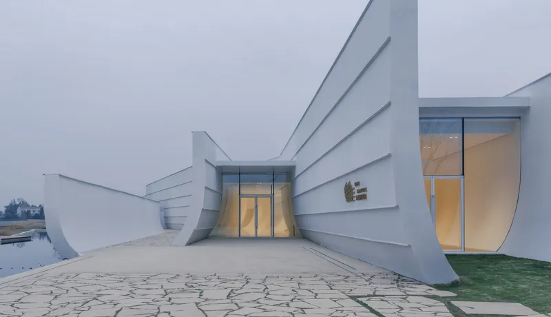 White facade with horizontal banding and illuminated arched openings at dusk above patterned paving