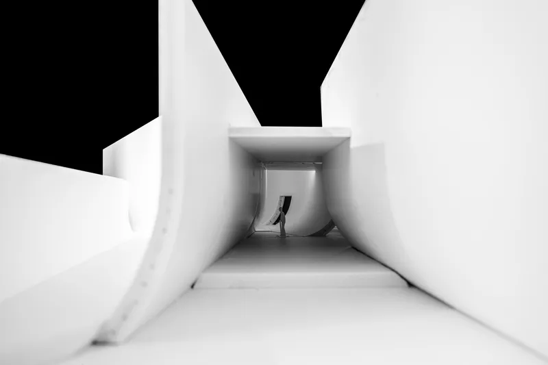 Interior view through a tapered corridor with a lone figure silhouetted against overhead skylight