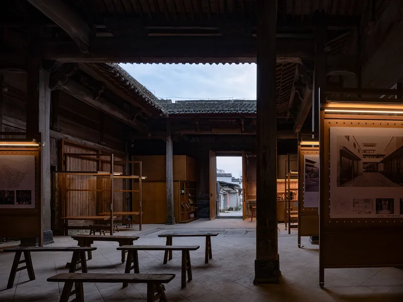 Open courtyard space framed by timber columns with wooden benches and illuminated display panels at dusk
