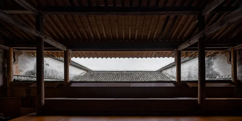 Framed view through timber structure to courtyard with tiled roof and white walls under overcast sky