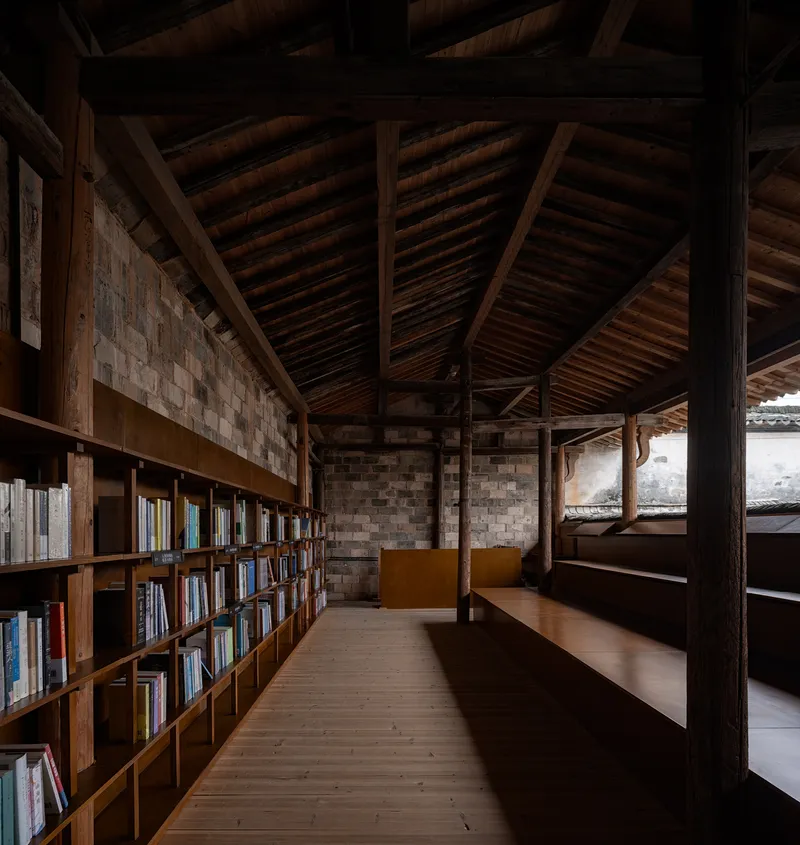 Library space with timber bookshelves along grey brick wall under exposed rafter ceiling and columns