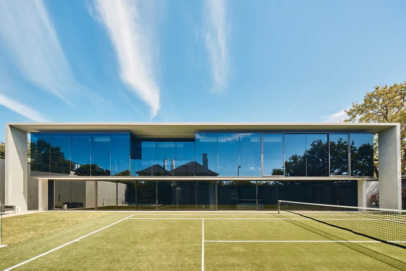 Rear glass facade overlooking a private grass tennis court under a clear morning sky
