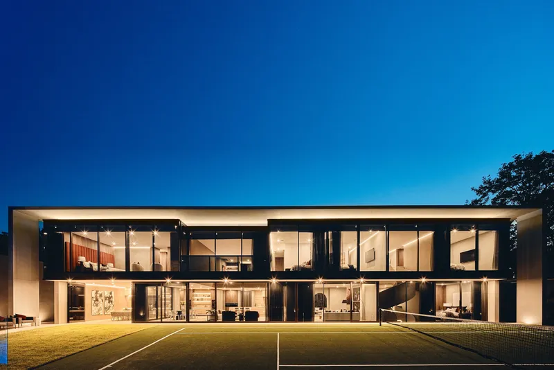 Rear elevation with full-height glazing across two floors overlooking the tennis court at twilight