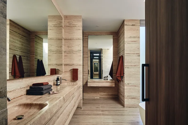 Bathroom vanity lined in travertine with mirrored wall reflecting the corridor beyond