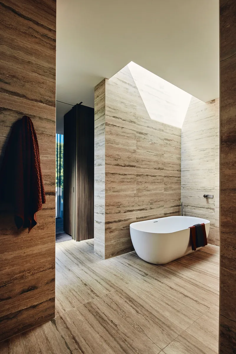 Freestanding white bathtub in travertine-clad bathroom with angled skylight overhead