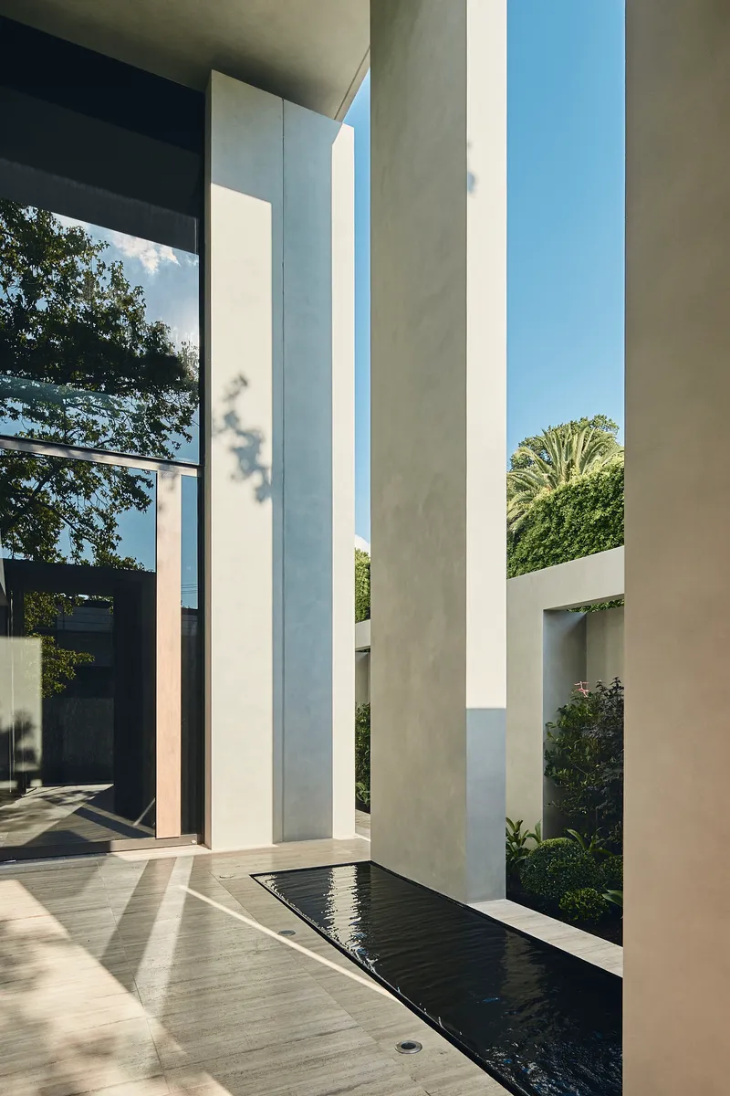 Concrete entry courtyard with tall vertical columns flanking a reflecting pool and planted beds