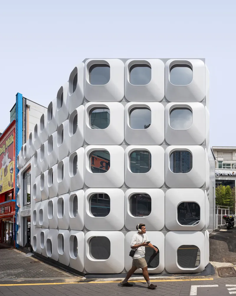 Corner facade showing stacked white modules with porthole windows as a person passes in front