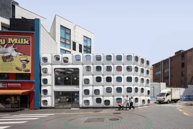 Streetscape showing the curved white facade with circular windows among neighboring buildings and passing vehicles