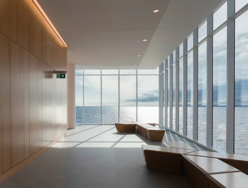 Interior corridor with full-height glazing overlooking the water and timber benches casting shadows on polished concrete