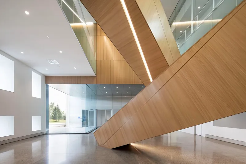 Double-height atrium with angular timber-clad ceiling planes and glass-enclosed mezzanine above polished concrete floor