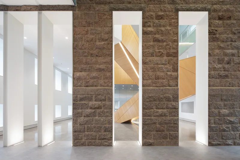 Tall vertical openings framing views of the timber staircase through a dark stone block wall