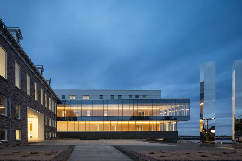 Illuminated glass facade extension alongside a brick heritage building at dusk