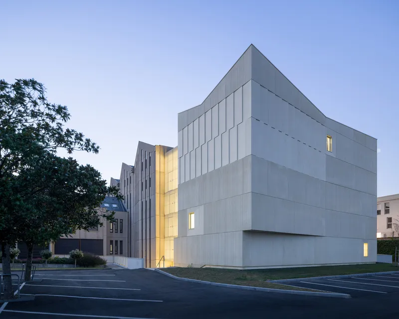 White paneled facade with vertical window slots illuminated at dusk beside a parking lot and tree
