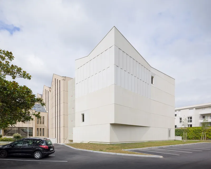 Corner view of the gabled volume with vertical panel cladding under overcast skies with parked car